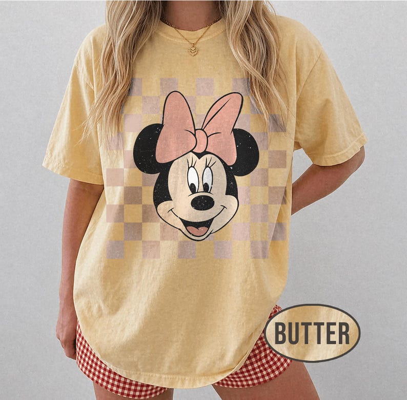 May include: Butter-colored t-shirt featuring a Minnie Mouse graphic with a pink bow and a checkered background. The word "BUTTER" is printed on the bottom right. The shirt has short sleeves and a relaxed fit.