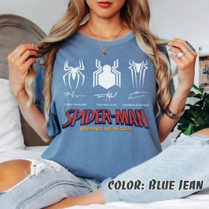 May include: A blue jean colored t-shirt featuring three white spider symbols and the text "SPIDER-MAN BRAND NEW DAY" in red and blue. The signatures of Tobey Maguire, Tom Holland, and Andrew Garfield are below the symbols.