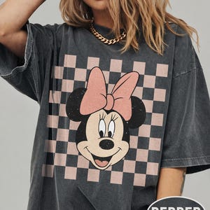 May include: Dark gray oversized t-shirt featuring a Minnie Mouse graphic. The graphic includes Minnie's face, a pink bow, and a checkered background in pink and black. The shirt has short sleeves and a relaxed fit.