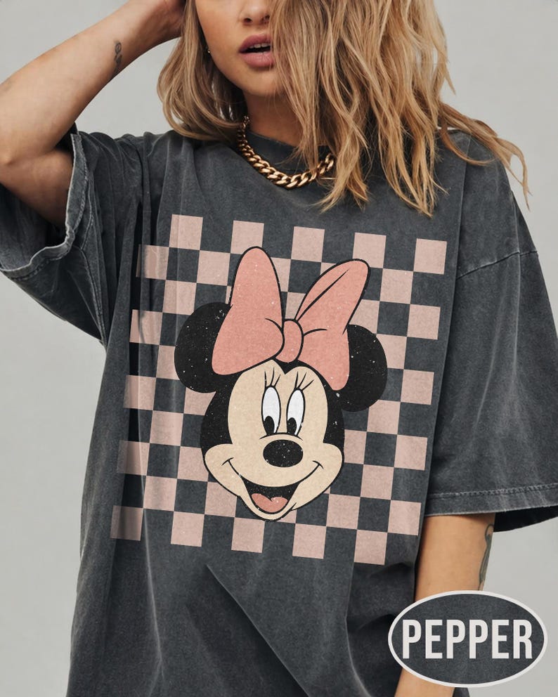 May include: Dark gray oversized t-shirt featuring a Minnie Mouse graphic. The graphic includes Minnie's face, a pink bow, and a checkered background in pink and black. The shirt has short sleeves and a relaxed fit.