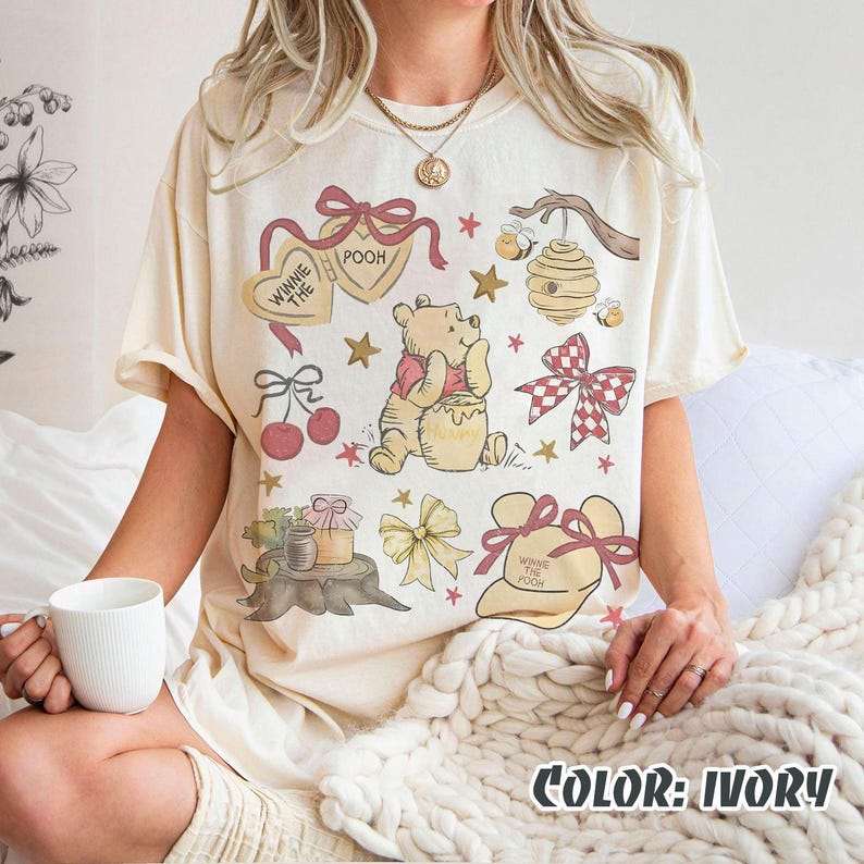 May include: Ivory t-shirt with a Winnie the Pooh design. The graphic includes Pooh Bear, a honey pot, bows, stars, cherries, and the text "Winnie the Pooh". The shirt has short sleeves and a loose fit.