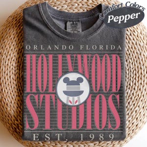 May include: A dark grey t-shirt with the text "Orlando Florida Hollywood Studios" in pink. The shirt features a Mickey Mouse icon and the text "EST. 1989". The shirt is folded on a woven surface.