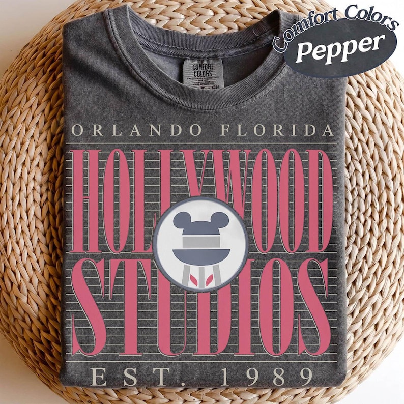 May include: A dark grey t-shirt with the text "Orlando Florida Hollywood Studios" in pink. The shirt features a Mickey Mouse icon and the text "EST. 1989". The shirt is folded on a woven surface.