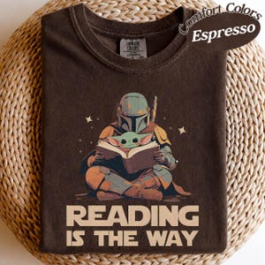 May include: Espresso-coloured t-shirt with a graphic of a Mandalorian character and Grogu reading a book. The text "READING IS THE WAY" is printed below the illustration. The shirt has a crew neck and short sleeves.