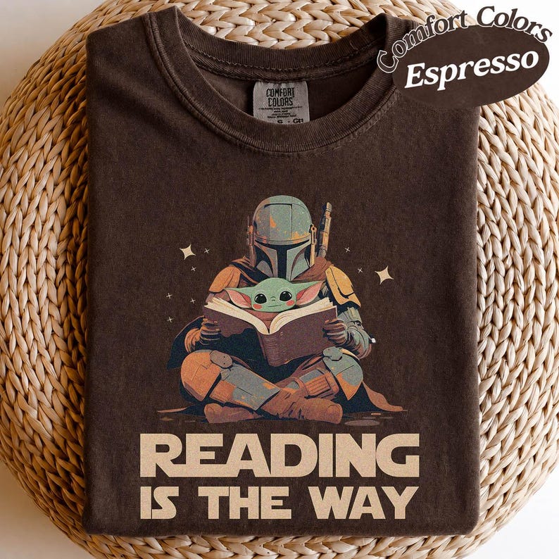May include: Espresso-coloured t-shirt with a graphic of a Mandalorian character and Grogu reading a book. The text "READING IS THE WAY" is printed below the illustration. The shirt has a crew neck and short sleeves.