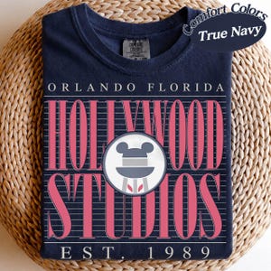 May include: A navy blue t-shirt featuring a pink graphic design for Hollywood Studios in Orlando, Florida. The design includes the words "Hollywood Studios", a Mickey Mouse logo, and "EST. 1989". The shirt is folded on a woven surface.