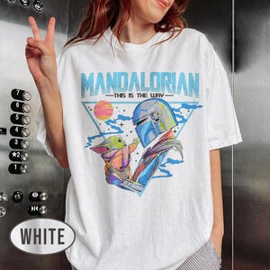 May include: White t-shirt featuring a colourful graphic of The Mandalorian and Grogu, with the text "MANDALORIAN - THIS IS THE WAY" in blue. The design includes a helmeted figure and a small green creature. The shirt has a casual, oversized fit.