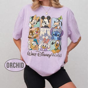 Op de afbeelding: Lichtpaars T-shirt met een rasterpatroon met Disney-personages zoals Mickey Mouse, Minnie Mouse, Donald Duck en Stitch. De woorden "Walt Disney World" staan eronder.
