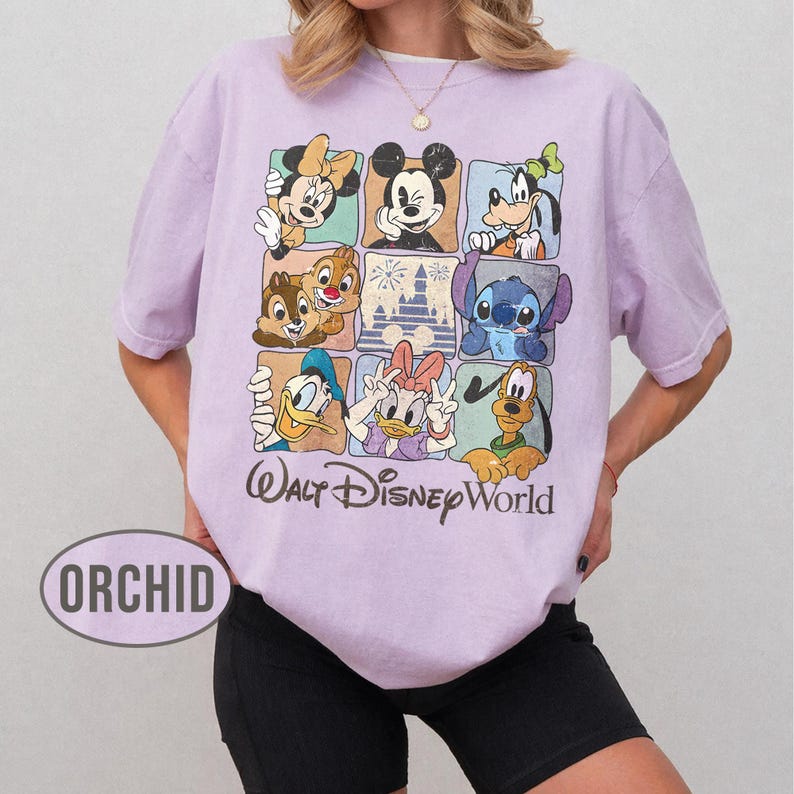 Op de afbeelding: Lichtpaars T-shirt met een rasterpatroon met Disney-personages zoals Mickey Mouse, Minnie Mouse, Donald Duck en Stitch. De woorden "Walt Disney World" staan eronder.