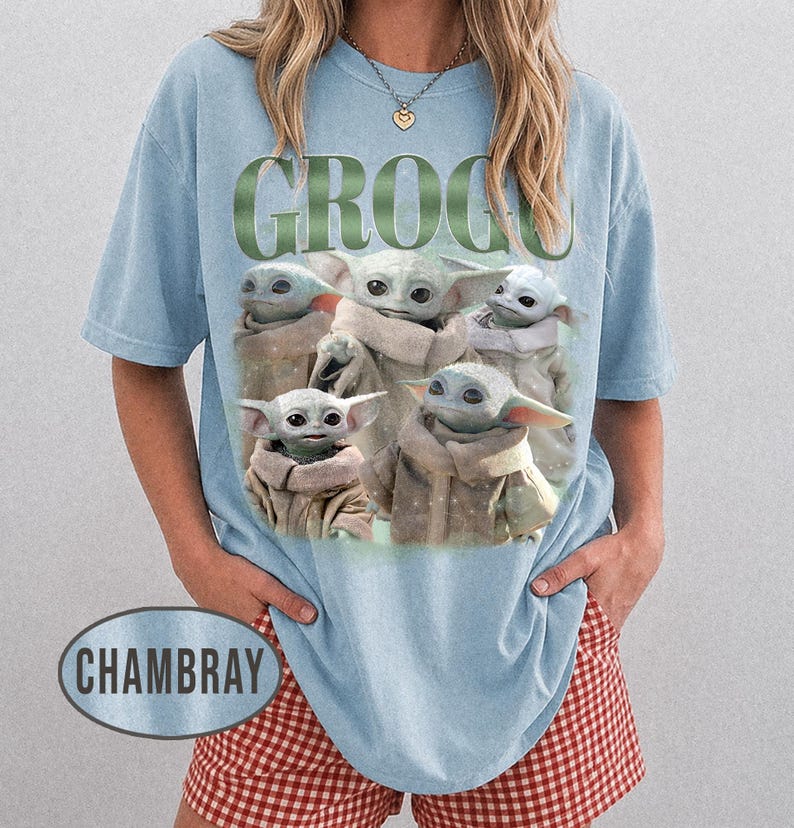 May include: Light blue t-shirt featuring a graphic of Grogu from The Mandalorian. The word "GROGU" is printed in green above the image of Grogu. The shirt is a light blue color.