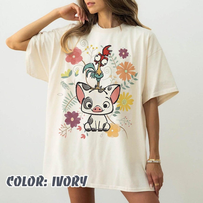 May include: Ivory-colored t-shirt featuring a cartoon pig with a rooster on its head, surrounded by colorful flowers and foliage. The text "COLOR: IVORY" is printed at the bottom. A casual, oversized fit.