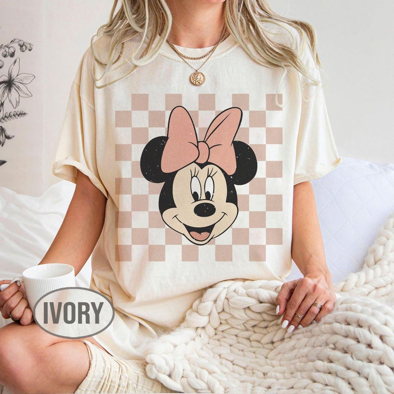 May include: Ivory-colored t-shirt featuring a Minnie Mouse graphic with a pink bow and a checkered background. The word "IVORY" is printed on a white mug. The shirt has short sleeves and a relaxed fit.