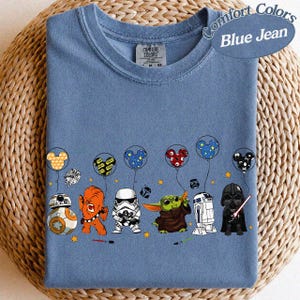 May include: Blue jean Comfort Colors t-shirt featuring Star Wars characters and Mickey Mouse-themed balloons. The design includes BB-8, Chewbacca, a Stormtrooper, Grogu, R2-D2, and Darth Vader. The shirt has a crew neck and short sleeves.