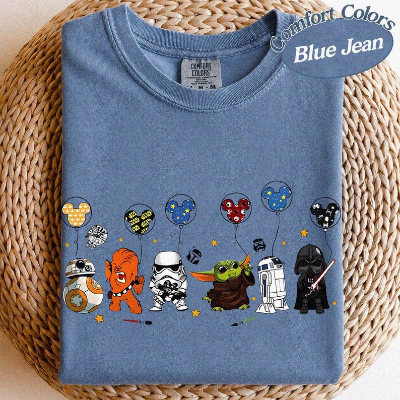 May include: Blue jean Comfort Colors t-shirt featuring Star Wars characters and Mickey Mouse-themed balloons. The design includes BB-8, Chewbacca, a Stormtrooper, Grogu, R2-D2, and Darth Vader. The shirt has a crew neck and short sleeves.