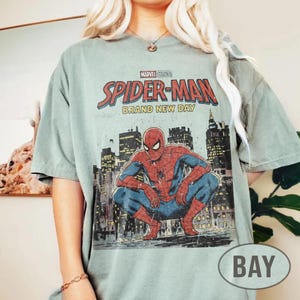 Może przedstawiać: Szałwiowa zielona koszulka z grafiką Spider-Mana kucającego przed panoramą miasta. Tekst "MARVEL STUDIOS SPIDER-MAN BRAND NEW DAY" jest wydrukowany u g&oacute;ry w kolorze czerwonym i białym.