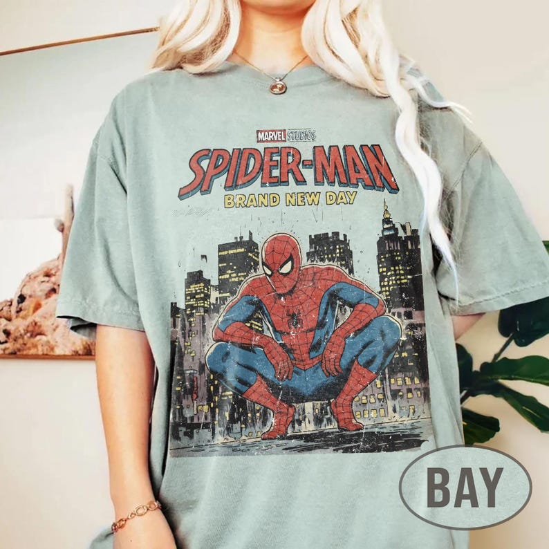 Może przedstawiać: Szałwiowa zielona koszulka z grafiką Spider-Mana kucającego przed panoramą miasta. Tekst "MARVEL STUDIOS SPIDER-MAN BRAND NEW DAY" jest wydrukowany u g&oacute;ry w kolorze czerwonym i białym.