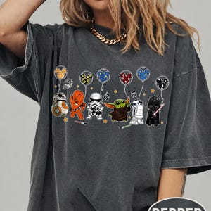 May include: Dark gray t-shirt featuring cartoon Star Wars characters holding balloons. The characters include BB-8, an orange Wookiee, a Stormtrooper, Grogu, R2-D2, and Darth Vader. The balloons have Mickey Mouse ears. The shirt has the word "PEPPER" printed on it.