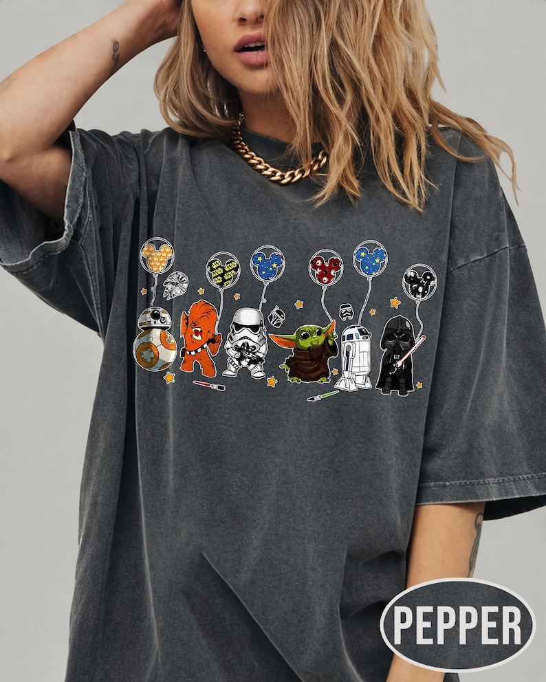 May include: Dark gray t-shirt featuring cartoon Star Wars characters holding balloons. The characters include BB-8, an orange Wookiee, a Stormtrooper, Grogu, R2-D2, and Darth Vader. The balloons have Mickey Mouse ears. The shirt has the word "PEPPER" printed on it.