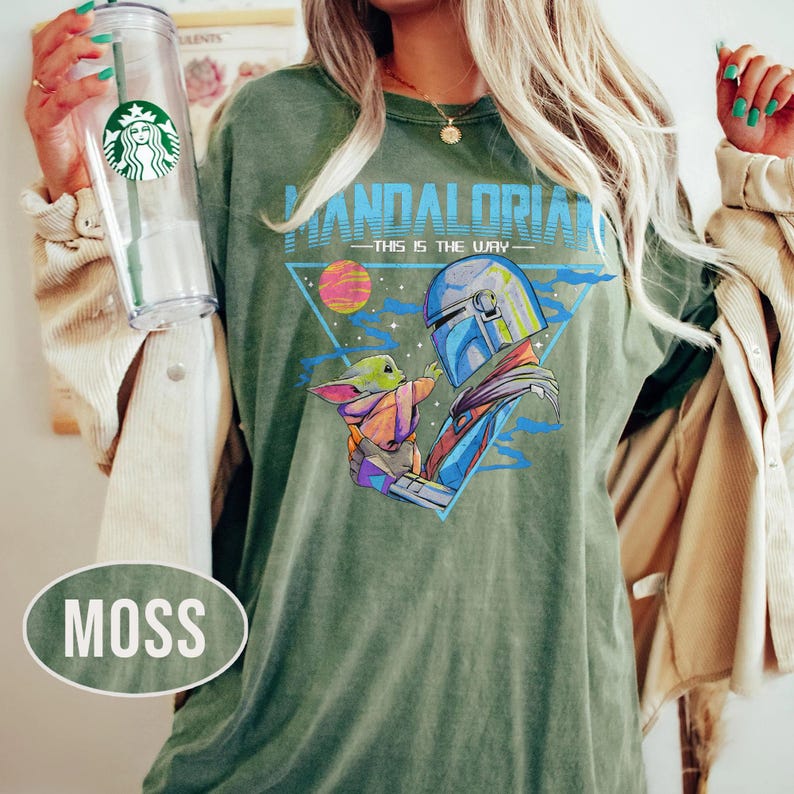 May include: Moss green t-shirt with a graphic of The Mandalorian and Grogu, and the text "MANDALORIAN - THIS IS THE WAY" in blue and white. The design includes a triangle, a planet, and a blue sky. The shirt has a relaxed fit.