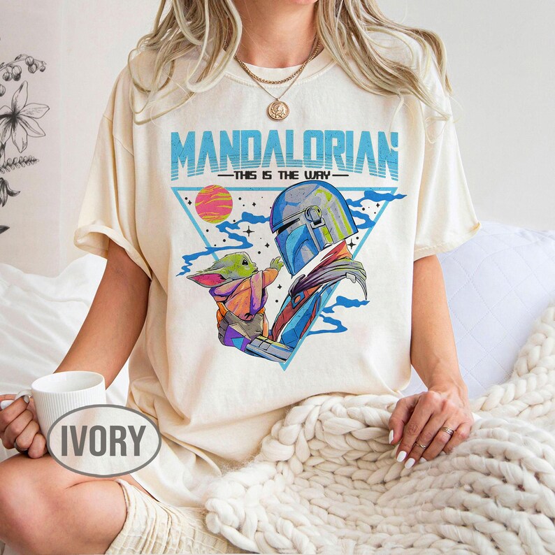 May include: Cream-coloured t-shirt with a graphic of the Mandalorian and Grogu, and the text "MANDALORIAN - THIS IS THE WAY" in blue. The design includes a triangle, a sunset, and a space scene. The shirt is worn by a person.