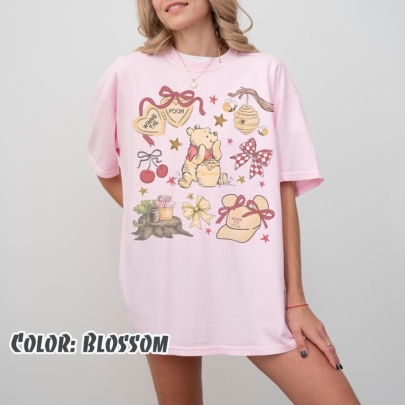May include: A pale pink t-shirt featuring a Winnie the Pooh design. The graphic includes Pooh Bear, honey pots, bows, and stars. The text "Winnie the Pooh" and "Pooh" are visible. The shirt's colour is described as "Blossom".