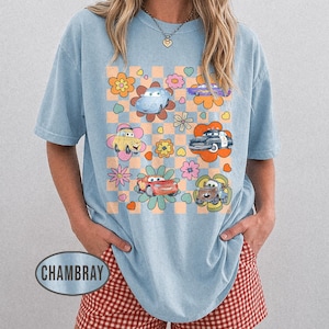 May include: Pale blue t-shirt with a graphic of cartoon cars and flowers on a checkered pattern. The shirt has short sleeves and a loose fit, suitable for everyday wear. The design is vibrant and playful.