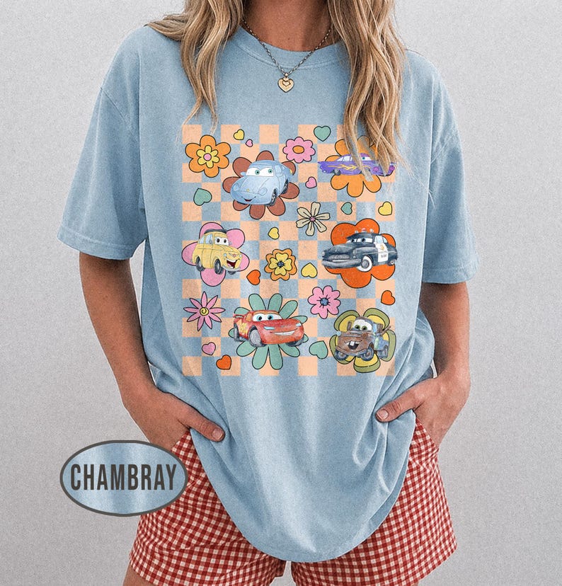 May include: Pale blue t-shirt with a graphic of cartoon cars and flowers on a checkered pattern. The shirt has short sleeves and a loose fit, suitable for everyday wear. The design is vibrant and playful.