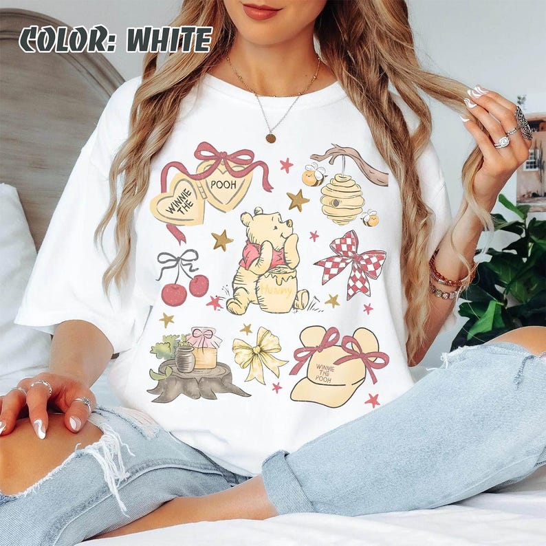 May include: White t-shirt with a colourful graphic of Winnie the Pooh, honey, bows, stars, and a beehive. The text "Winnie the Pooh" is also present. The shirt has a relaxed fit.
