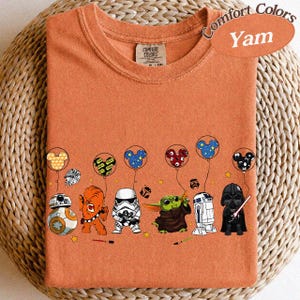 May include: Orange Comfort Colors t-shirt with a graphic of Star Wars characters holding Mickey Mouse balloons. The characters include BB-8, Chewbacca, a Stormtrooper, Grogu, R2-D2, and Darth Vader. The shirt is labeled "Yam".