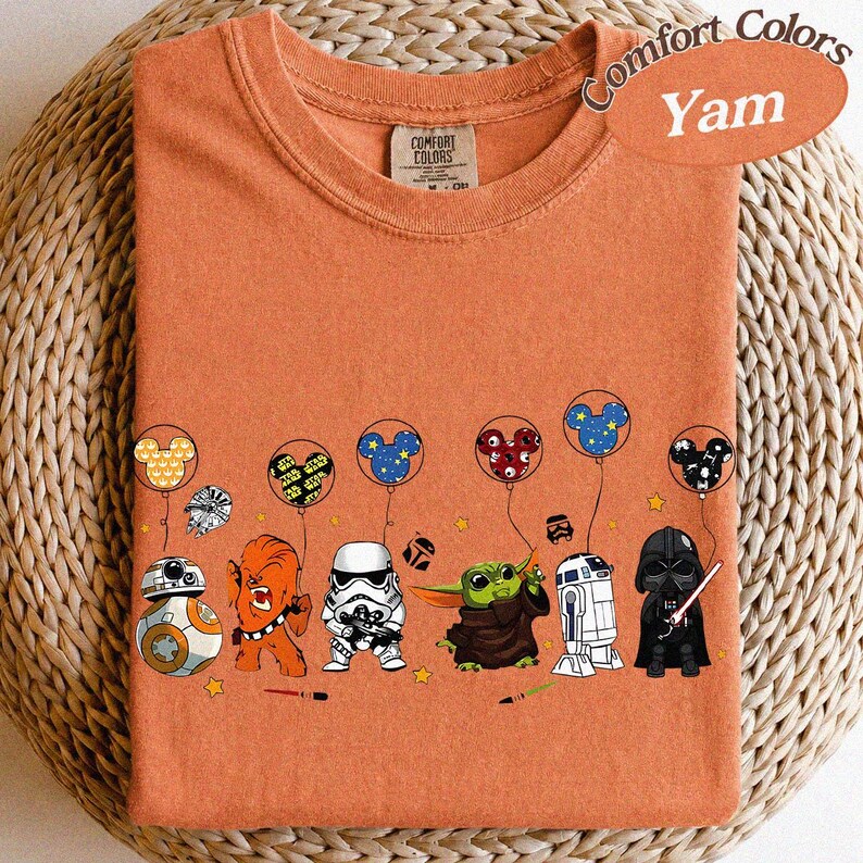 May include: Orange Comfort Colors t-shirt with a graphic of Star Wars characters holding Mickey Mouse balloons. The characters include BB-8, Chewbacca, a Stormtrooper, Grogu, R2-D2, and Darth Vader. The shirt is labeled "Yam".