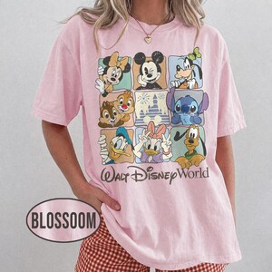 Op de afbeelding: Lichtroze T-shirt met een rasterpatroon van Disney-personages, waaronder Mickey Mouse, Minnie Mouse en Donald Duck. De tekst "Walt Disney World" staat eronder gedrukt. Het T-shirt heeft korte mouwen.