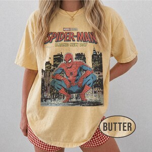 Może przedstawiać: Maślana koszulka z grafiką Spider-Mana. Projekt zawiera napis "Marvel Studios Spider-Man Brand New Day" nad ilustracją Spider-Mana kucającego przed panoramą miasta. Koszulka ma lekko wyblakły, zużyty wygląd.