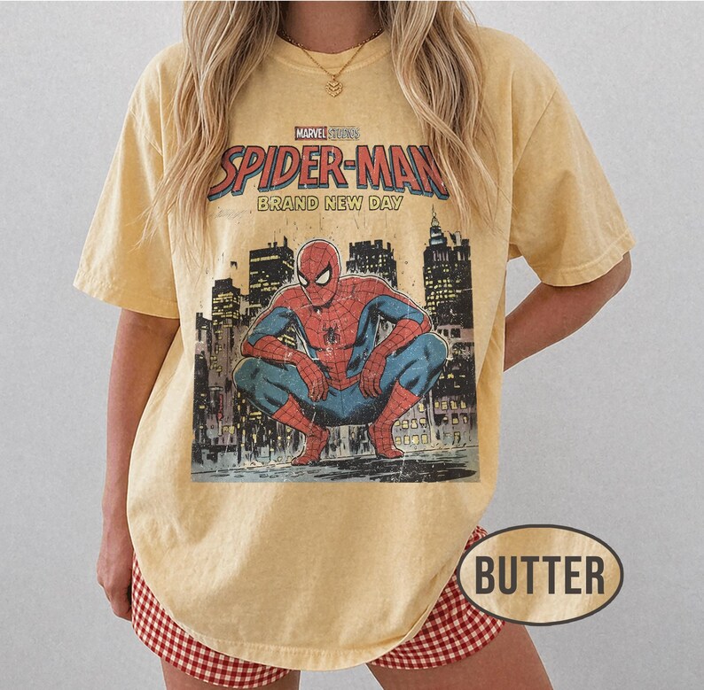 Może przedstawiać: Maślana koszulka z grafiką Spider-Mana. Projekt zawiera napis "Marvel Studios Spider-Man Brand New Day" nad ilustracją Spider-Mana kucającego przed panoramą miasta. Koszulka ma lekko wyblakły, zużyty wygląd.