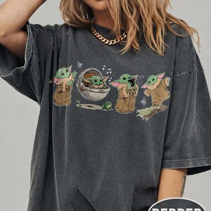 May include: Dark grey t-shirt with a graphic of a character from a popular science fiction series. The design shows the character in various poses, with a frog and musical notes. The shirt has a relaxed fit and the word "PEPPER" is printed on the bottom right.