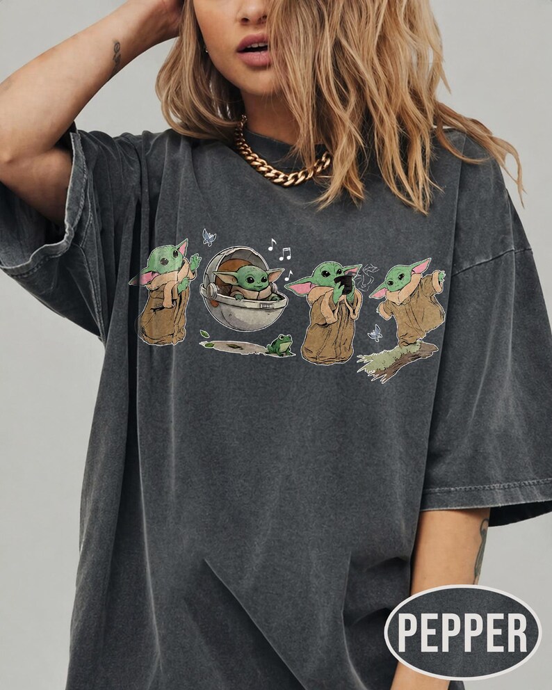 May include: Dark grey t-shirt with a graphic of a character from a popular science fiction series. The design shows the character in various poses, with a frog and musical notes. The shirt has a relaxed fit and the word "PEPPER" is printed on the bottom right.