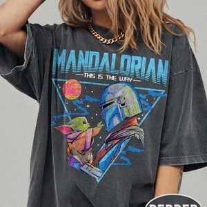 May include: Dark grey t-shirt with a graphic of The Mandalorian and Grogu, and the text "MANDALORIAN - THIS IS THE WAY" in blue. The design includes a triangle, a planet, and a space scene. The shirt has short sleeves.