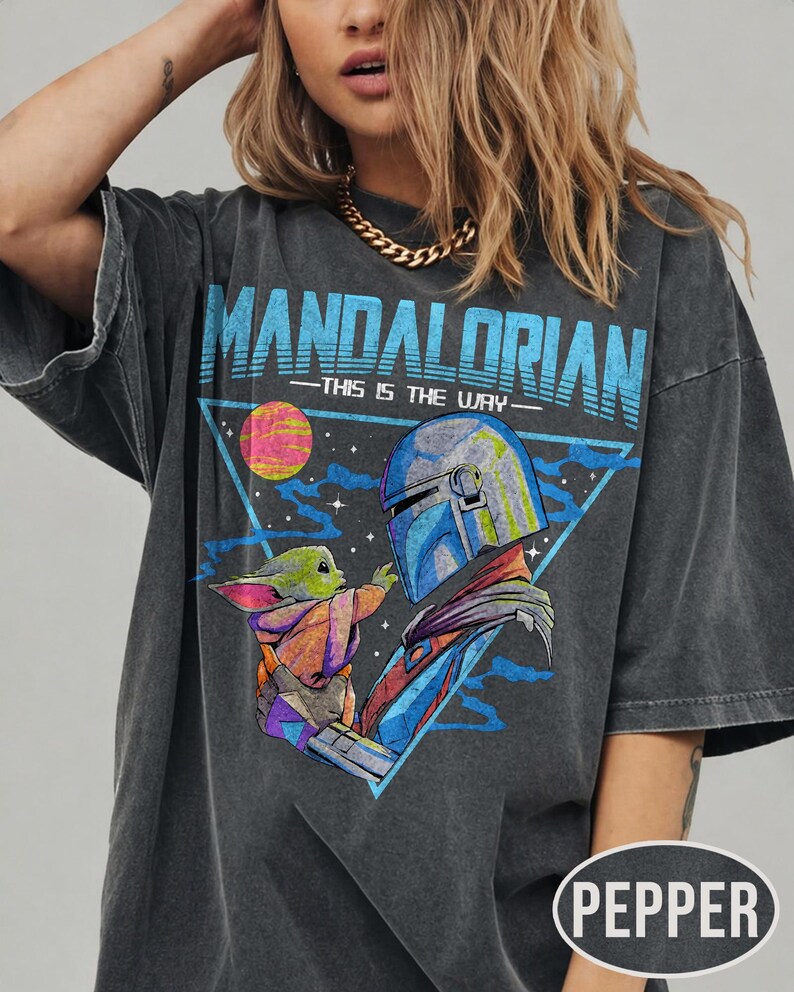 May include: Dark grey t-shirt with a graphic of The Mandalorian and Grogu, and the text "MANDALORIAN - THIS IS THE WAY" in blue. The design includes a triangle, a planet, and a space scene. The shirt has short sleeves.