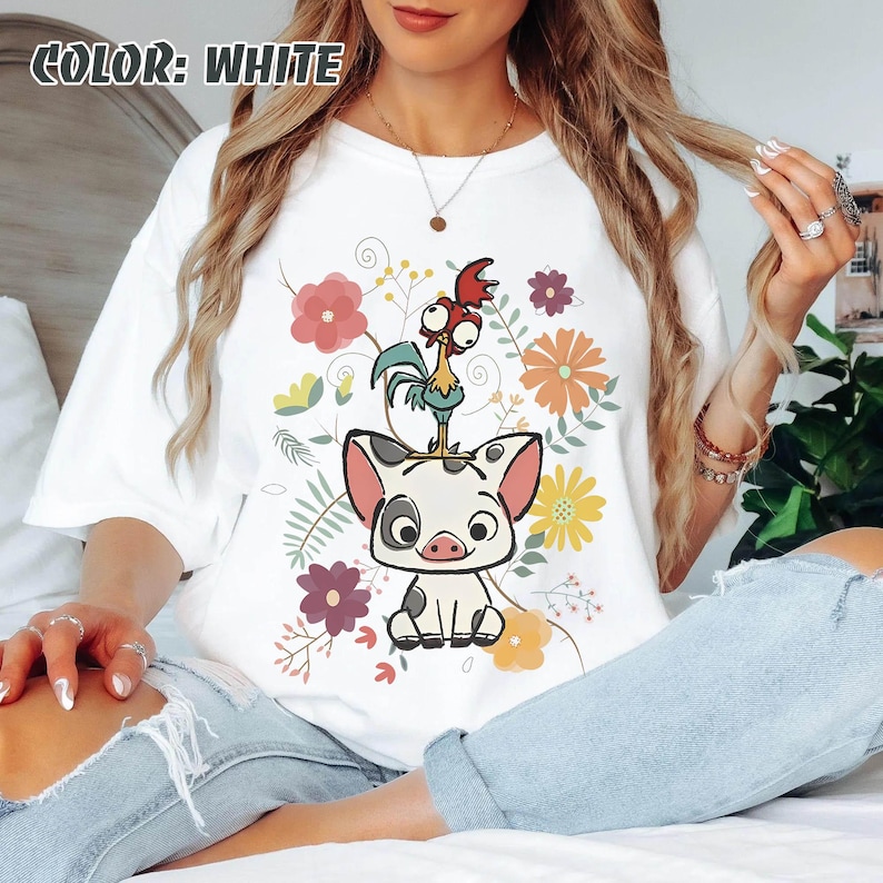 Puede incluir: Camiseta blanca con un dise&ntilde;o de cerdo y gallo de dibujos animados, sobre un fondo de flores y hojas coloridas. El texto "COLOR: WHITE" es visible. El dise&ntilde;o incorpora tonos rosas, naranjas, amarillos y verdes.