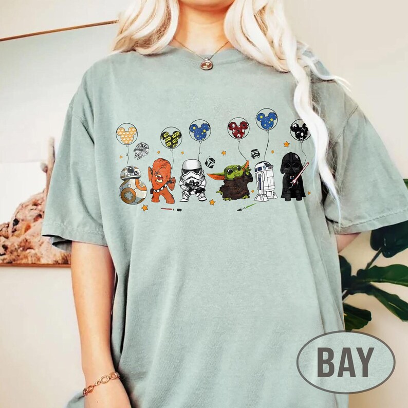 May include: A light green t-shirt featuring cartoon characters from Star Wars, each holding a Mickey Mouse-shaped balloon. Characters include BB-8, an orange alien, a Stormtrooper, Grogu, R2-D2, and Darth Vader. The shirt has a relaxed fit.