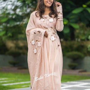 May include: A light peach-colored long-sleeved dress featuring a square neckline and a matching draped shawl. The garment is adorned with hand-painted brown and white floral motifs across the bodice and shawl. The text Bahe Kurtiz is visible.