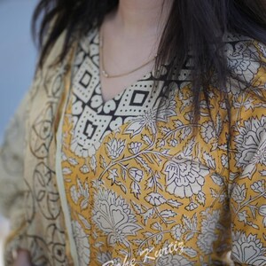 May include: A yellow and white floral print kurta with a black and white geometric print on the neckline and cuffs. The kurta is made of cotton and has a V-neckline. The model is wearing a green and silver bracelet.