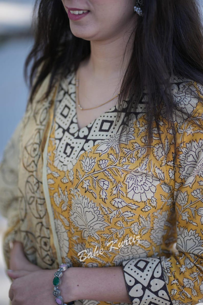 May include: A yellow and white floral print kurta with a black and white geometric print on the neckline and cuffs. The kurta is made of cotton and has a V-neckline. The model is wearing a green and silver bracelet.