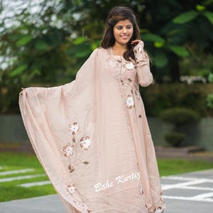 May include: A light peach-colored long-sleeved kurta dress featuring a matching sheer dupatta with hand-painted white floral designs. The garment includes the text Bahe Kurtiz printed in white on the lower section of the fabric.
