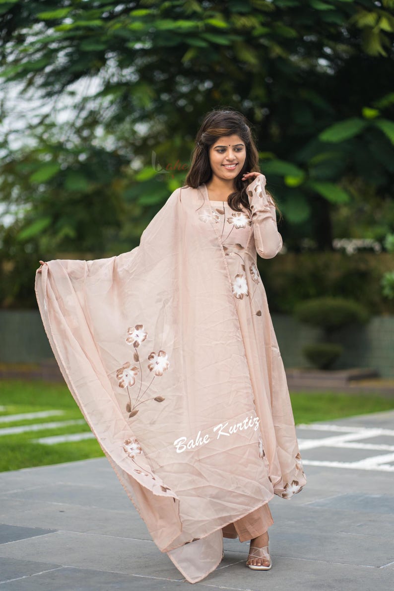 May include: A light peach-colored long-sleeved kurta dress featuring a matching sheer dupatta with hand-painted white floral designs. The garment includes the text Bahe Kurtiz printed in white on the lower section of the fabric.