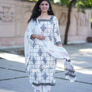 May include: A white cotton kurta with a blue and white geometric print and a matching white cotton palazzo pant with a red and white geometric print. The kurta has a square neckline and three-quarter sleeves. The model is wearing a white sheer dupatta with a blue and white border.