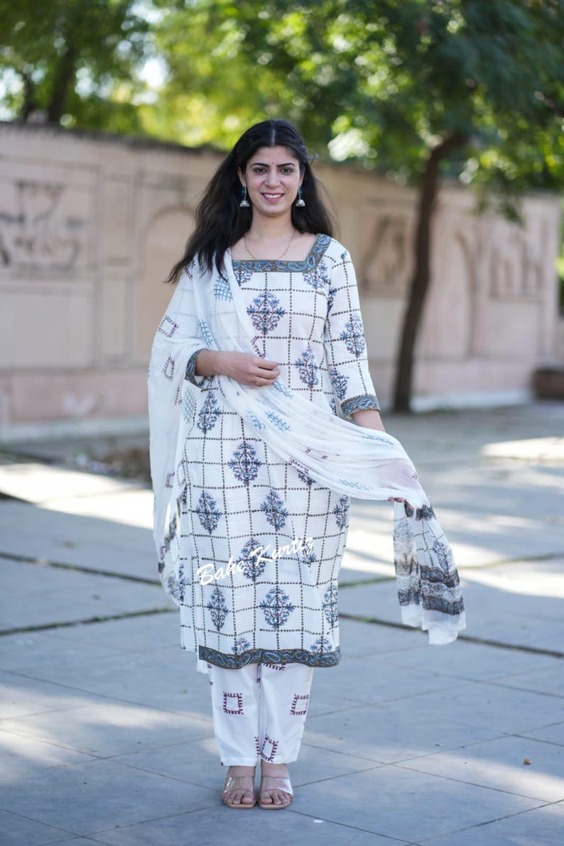 May include: A white cotton kurta with a blue and white geometric print and a matching white cotton palazzo pant with a red and white geometric print. The kurta has a square neckline and three-quarter sleeves. The model is wearing a white sheer dupatta with a blue and white border.