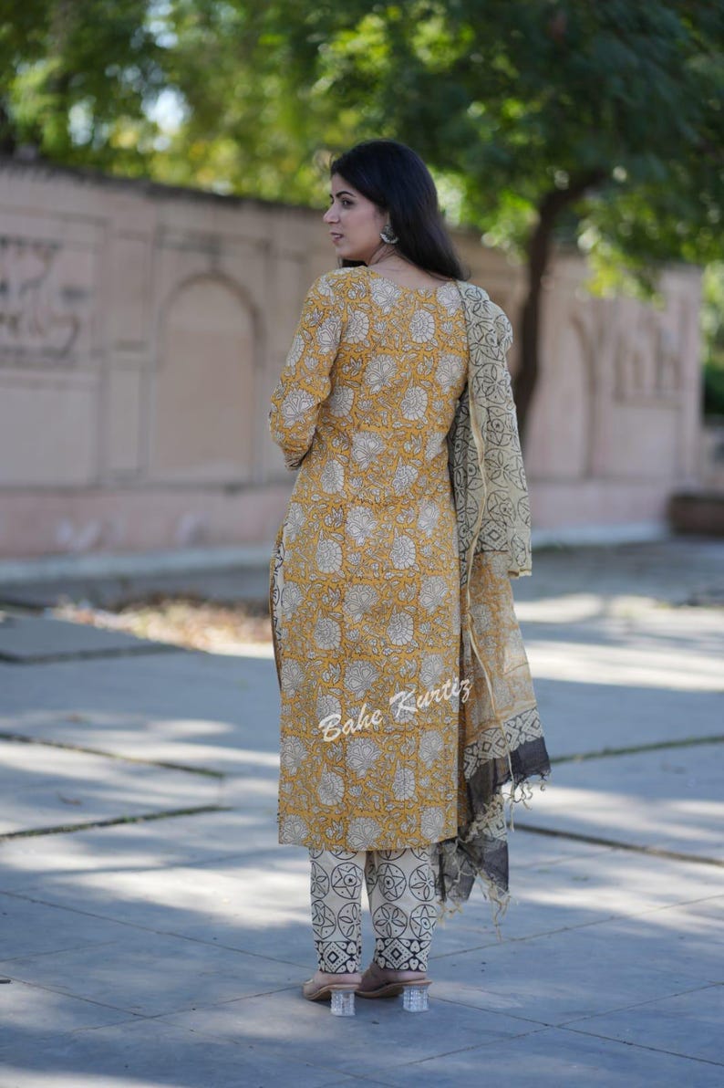 May include: A yellow and white floral print kurta with matching pants and a cream and black dupatta. The kurta has a round neckline and three-quarter sleeves. The pants have a straight leg and the dupatta is a sheer fabric with a black border.