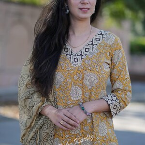 May include: A woman wearing a yellow and white floral printed kurta with a brown and white geometric pattern on the neckline and sleeves. She is also wearing a matching dupatta with a similar pattern. The kurta is a traditional Indian garment.