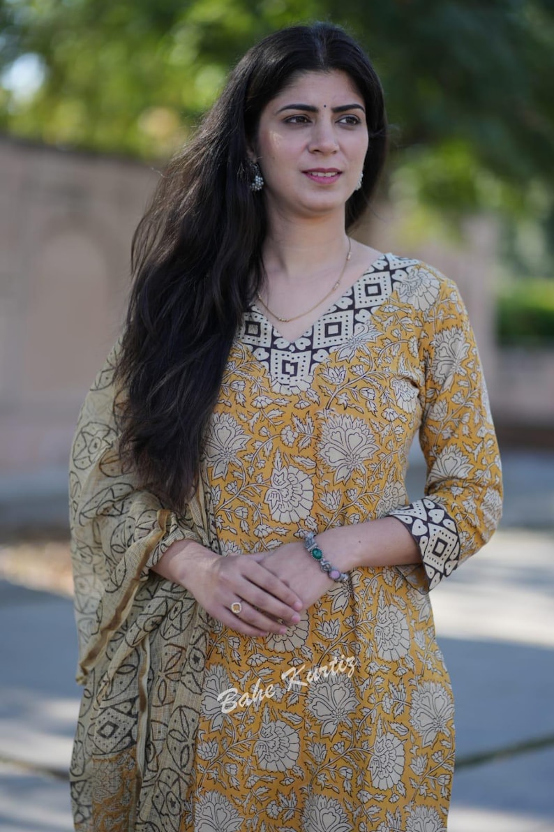 May include: A woman wearing a yellow and white floral printed kurta with a brown and white geometric pattern on the neckline and sleeves. She is also wearing a matching dupatta with a similar pattern. The kurta is a traditional Indian garment.