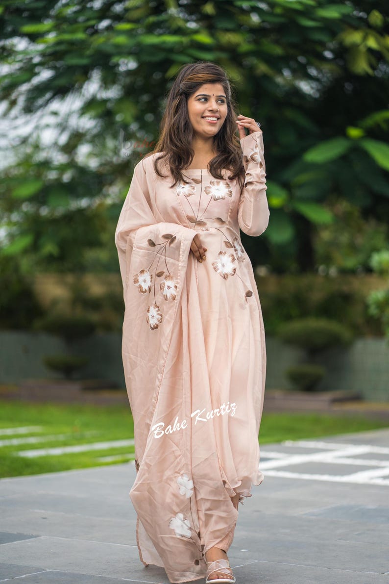 May include: A light peach-colored long-sleeved dress featuring a square neckline and a matching draped shawl. The garment is adorned with hand-painted brown and white floral motifs across the bodice and shawl. The text Bahe Kurtiz is visible.